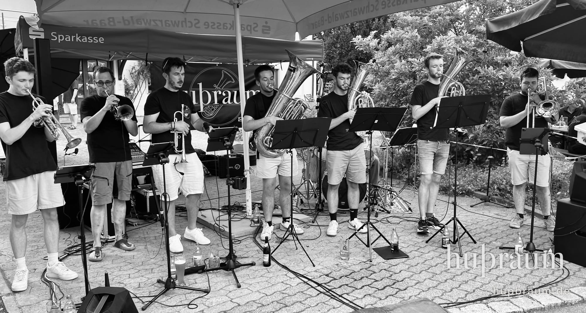 huþraum beim Auftritt: sieben Musizierende in schwarzen Shirts unter einem Sonnenschirm, Trompeten, Tuben und Posaune, im Hintergrund das Bandbanner.